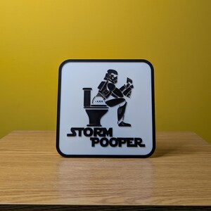 May include: A white square sign with a black border. The sign features a black silhouette of a Stormtrooper sitting on a toilet. The text "Storm Pooper" is printed in black below the silhouette.