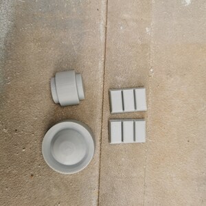 May include: Four gray 3D printed objects: a round disc, a cylinder, and two rectangular blocks with raised lines.