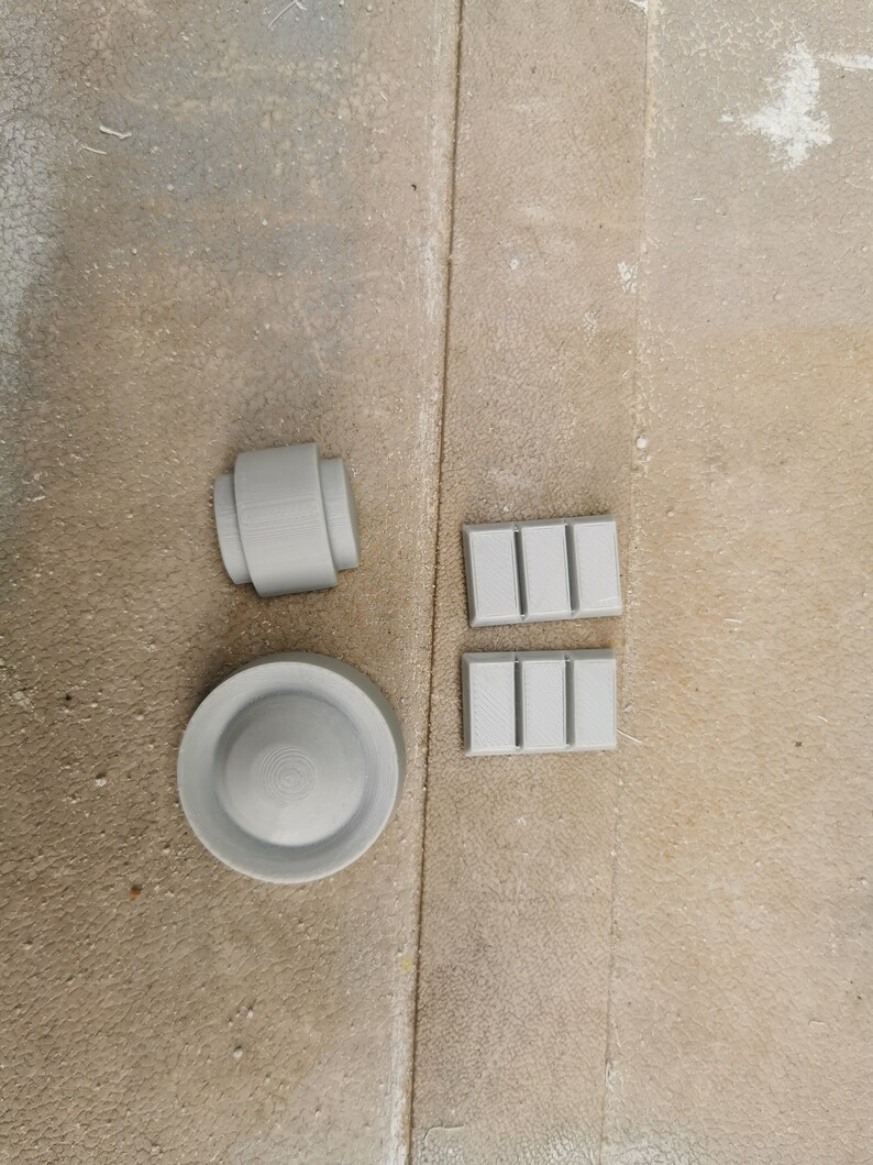 May include: Four gray 3D printed objects: a round disc, a cylinder, and two rectangular blocks with raised lines.