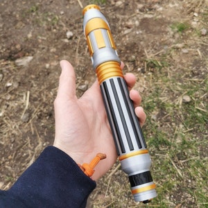 May include: A silver and gold toy prop resembling a futuristic pen or tool. The prop has a black and silver striped design with gold accents.