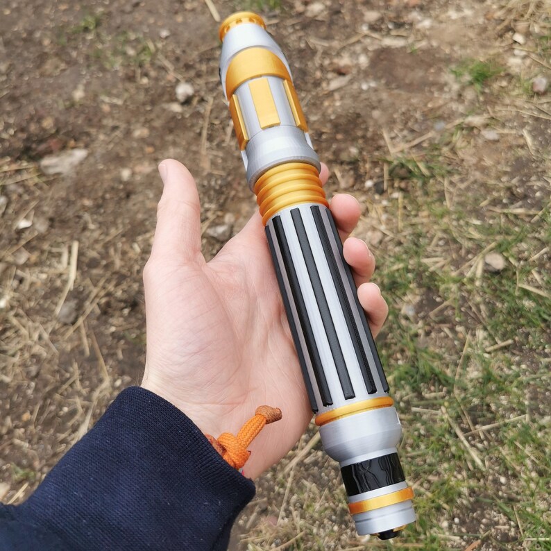 May include: A silver and gold toy prop resembling a futuristic pen or tool. The prop has a black and silver striped design with gold accents.