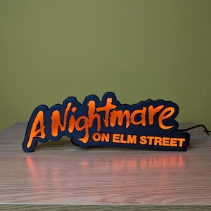 May include: A black and orange LED sign with the text "A Nightmare on Elm Street". The sign has a black base and the text is in a stylized orange font. The sign is lit up and plugged into a power source.