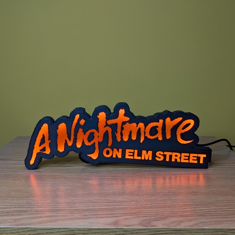 May include: A black and orange LED sign with the text "A Nightmare on Elm Street". The sign has a black base and the text is in a stylized orange font. The sign is lit up and plugged into a power source.
