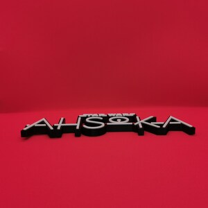 May include: Black and white 3D-printed sign with the text "STAR WARS AHSOKA" on a red background. The letters are black with white outlines, creating a layered effect. The sign is a decorative item.