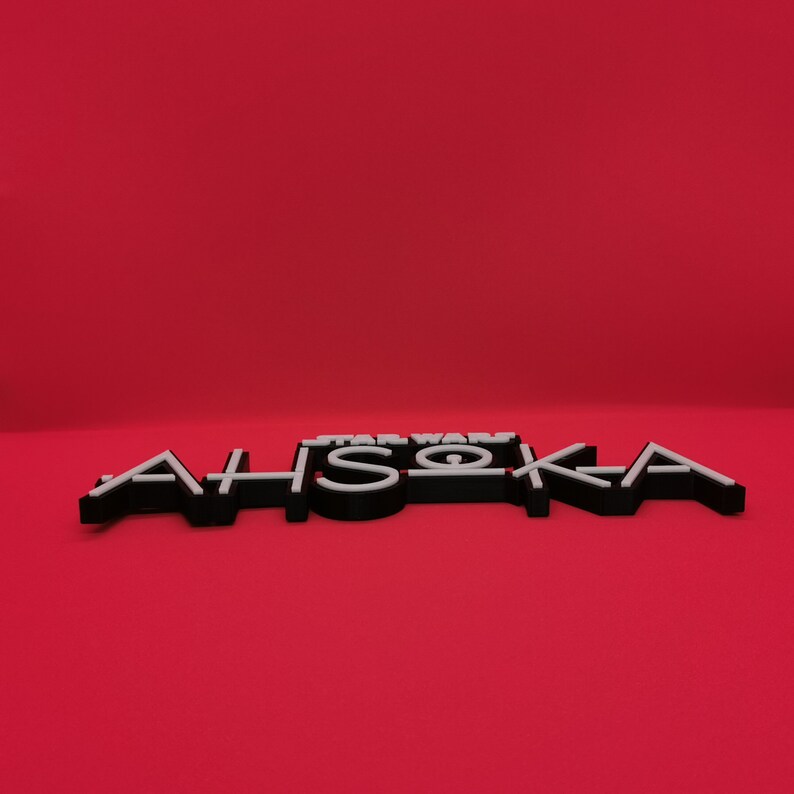 May include: Black and white 3D-printed sign with the text "STAR WARS AHSOKA" on a red background. The letters are black with white outlines, creating a layered effect. The sign is a decorative item.