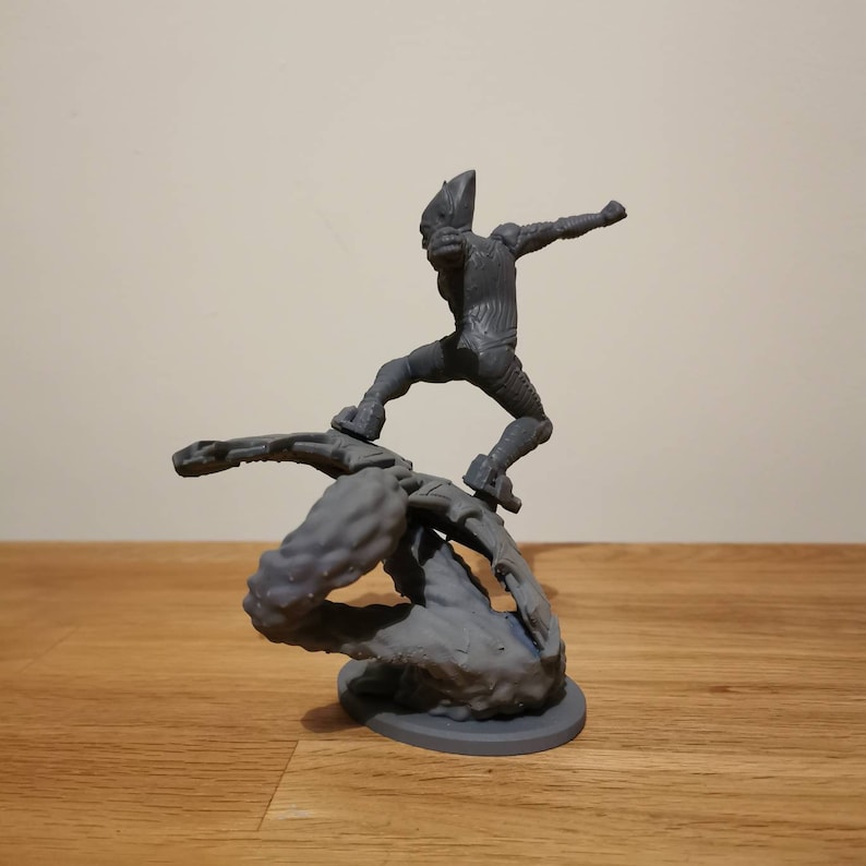 May include: A gray 3D printed figurine of a superhero standing on a web-like platform. The superhero is wearing a suit with a textured design and has a mask covering their face. The platform is made of a web-like material and is attached to a base.