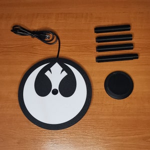 May include: A black and white Rebel Alliance symbol on a round, black charging pad with a USB cable. The charging pad is on a wooden surface with four black charging stands.