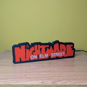 May include: A black and red sign with the text "A Nightmare on Elm Street" in a stylized font. The sign is on a wooden surface with a yellow background.