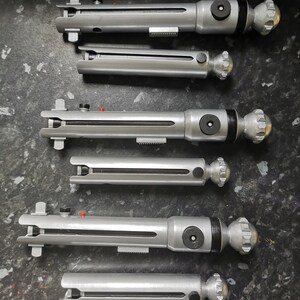 May include: Six silver lightsaber hilts with black accents and a silver knob on the end. The hilts are arranged vertically in a row.