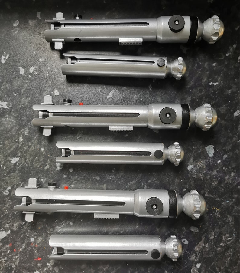May include: Six silver lightsaber hilts with black accents and a silver knob on the end. The hilts are arranged vertically in a row.