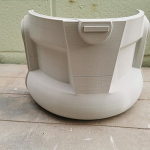 May include: A white plastic helmet with a smooth, rounded surface and a rectangular detail on the front. The helmet is sitting on a wooden surface.