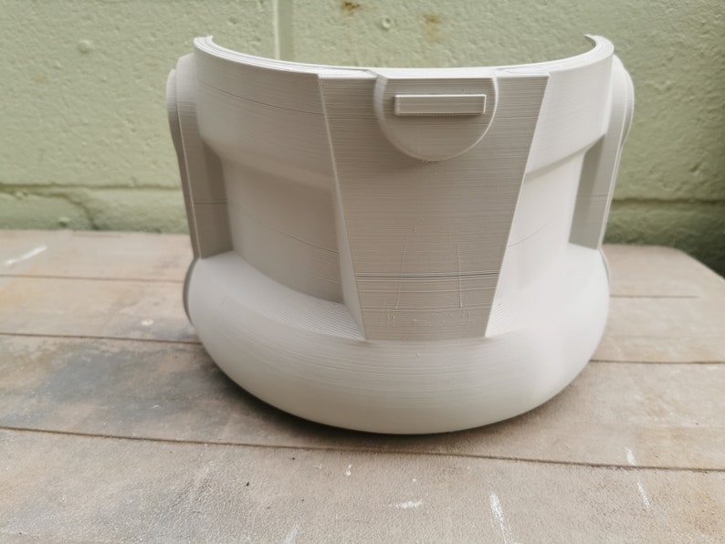 May include: A white plastic helmet with a smooth, rounded surface and a rectangular detail on the front. The helmet is sitting on a wooden surface.