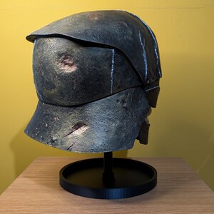 May include: A weathered, dark gray helmet with a distressed finish. The helmet has a textured surface and appears to be made of metal. It is displayed on a black stand.