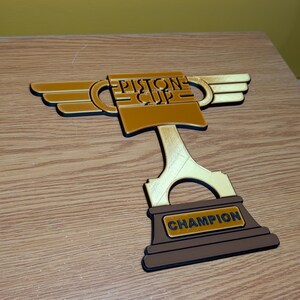May include: A gold-colored trophy with a piston design and the words "PISTONE CUP" on the cup. The base of the trophy is brown and says "CHAMPION".