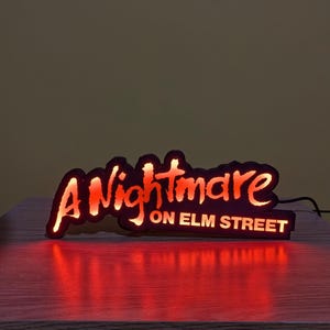 May include: Illuminated sign with the text "A Nightmare on Elm Street" in red and orange. The sign is black with a glowing effect, casting a red reflection on the surface below. A horror movie-themed decorative item.