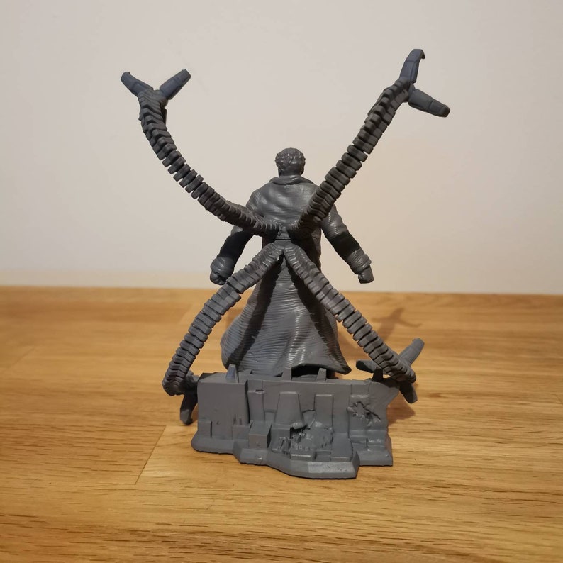 May include: A gray plastic action figure of Doctor Octopus from the Marvel universe. The figure is posed with its four mechanical arms extended outward. The figure is standing on a gray plastic base that resembles a destroyed building.