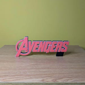 May include: A red and black Avengers logo, a decorative item, is displayed on a wooden surface. The word "Avengers" is in a bold, stylized font with a black outline. The logo is a vibrant red color.
