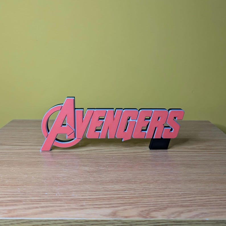 May include: A red and black Avengers logo, a decorative item, is displayed on a wooden surface. The word "Avengers" is in a bold, stylized font with a black outline. The logo is a vibrant red color.