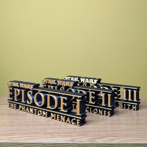May include: Three black and gold Star Wars Episode titles. The first title is "Episode I The Phantom Menace", the second is "Episode II The Clones", and the third is "Episode III Sith".