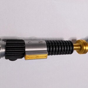 May include: A detailed replica lightsaber, featuring a silver hilt with black ribbed grip, gold accents, and a clear end cap. The design includes a yellow rectangular detail and a silver pommel. This collectible item is a detailed representation of a lightsaber.