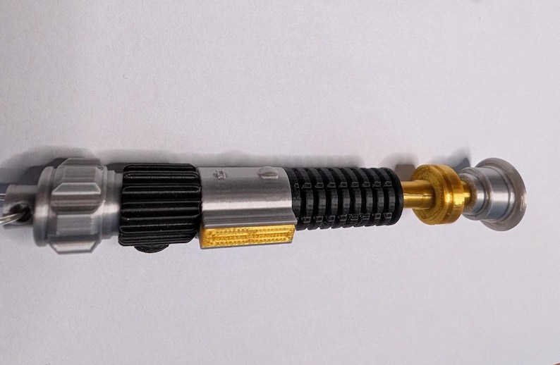 May include: A detailed replica lightsaber, featuring a silver hilt with black ribbed grip, gold accents, and a clear end cap. The design includes a yellow rectangular detail and a silver pommel. This collectible item is a detailed representation of a lightsaber.