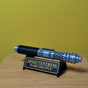 May include: A black and silver lightsaber with a honeycomb design on the barrel. The lightsaber is on a black stand with the text "Asajj Ventress Dark Disciple" in gold lettering.