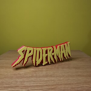 May include: A 3D printed word mark of "SPIDER-MAN" in yellow letters with red outlines. The letters are stylized and have a slightly jagged appearance.