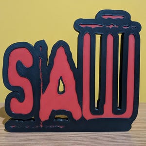 May include: A black and red wooden sign with the word "SAW" in a stylized font. The "A" is larger than the other letters and has a jagged edge. The sign is shaped like a trash can.