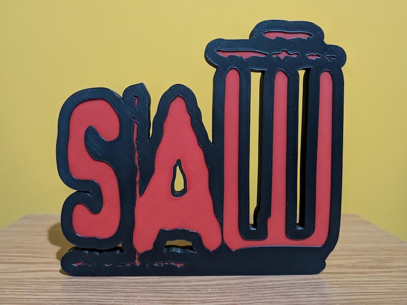 May include: A black and red wooden sign with the word "SAW" in a stylized font. The "A" is larger than the other letters and has a jagged edge. The sign is shaped like a trash can.