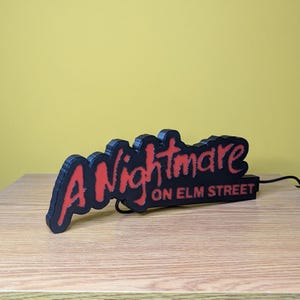 May include: A black and red sign that reads "A Nightmare on Elm Street." The sign is 3D printed and sits on a wooden surface. The text is in a stylized font, with the word "Nightmare" in red.