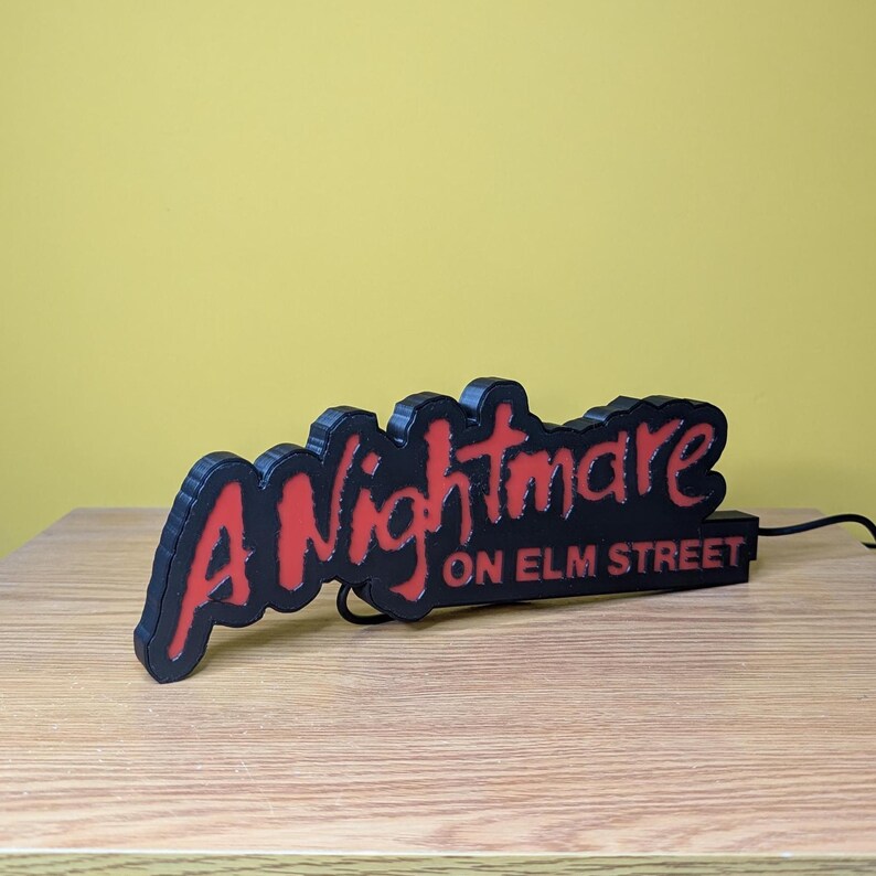 May include: A black and red sign that reads "A Nightmare on Elm Street." The sign is 3D printed and sits on a wooden surface. The text is in a stylized font, with the word "Nightmare" in red.