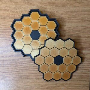 May include: Two black and gold honeycomb-shaped coasters. The larger coaster has a black center and the smaller coaster has a black center.