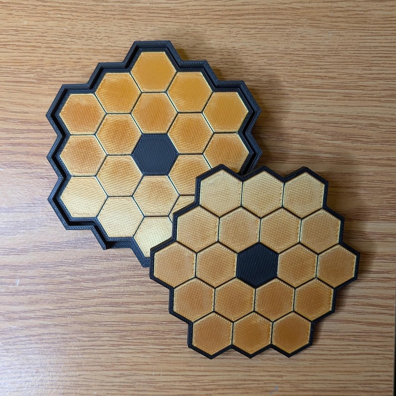 May include: Two black and gold honeycomb-shaped coasters. The larger coaster has a black center and the smaller coaster has a black center.