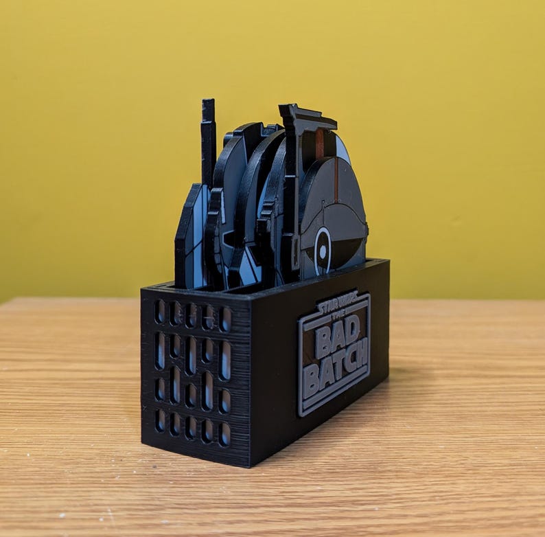 May include: A black and gray Star Wars: The Bad Batch themed coaster set with a black stand. The coasters feature various characters and vehicles from the show.