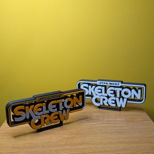 May include: Two black and white 3D printed signs with the text "Star Wars Skeleton Crew" in a retro font. The top sign has white text and the bottom sign has gold text.