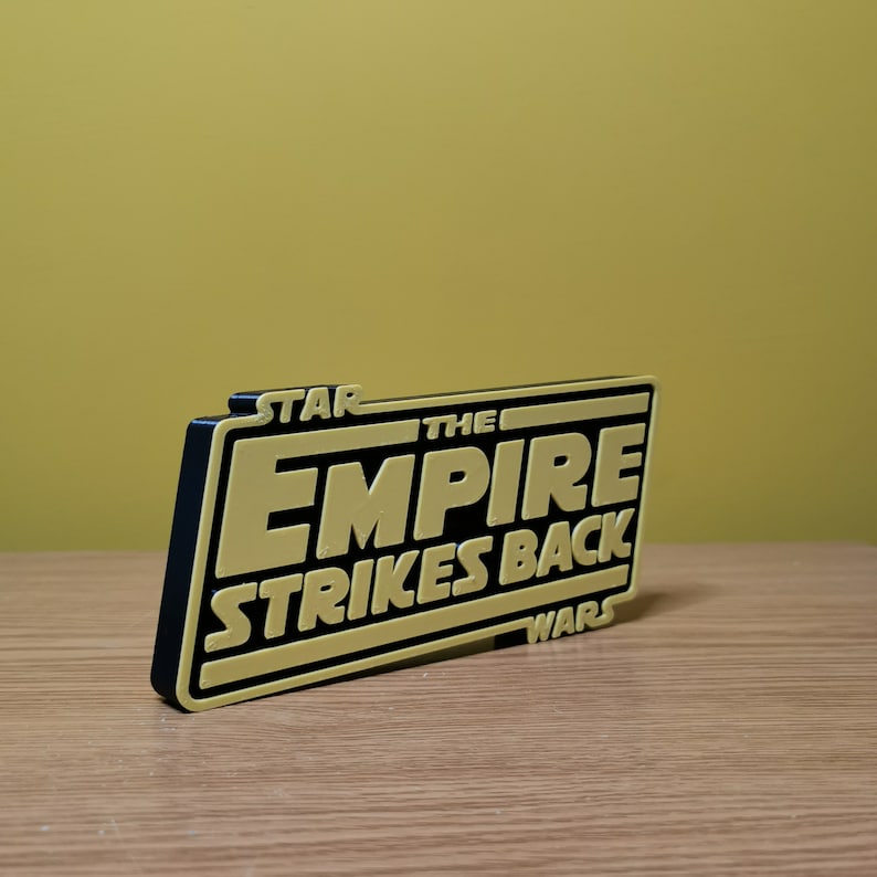 May include: A black and gold 3D printed sign with the text "STAR WARS THE EMPIRE STRIKES BACK".