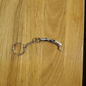 Op de afbeelding: Een zilveren sleutelhanger met een zwart en zilveren lightsaber-bedel. De sleutelhanger heeft een zilveren ring en een zilveren ketting.