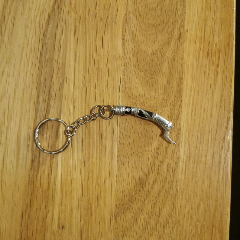 Op de afbeelding: Een zilveren sleutelhanger met een zwart en zilveren lightsaber-bedel. De sleutelhanger heeft een zilveren ring en een zilveren ketting.