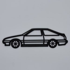 May include: Black outline of a classic sports car, featuring a two-door coupe design. The car has detailed wheels and a sleek profile, set against a plain white background. The design is minimalist and graphic.
