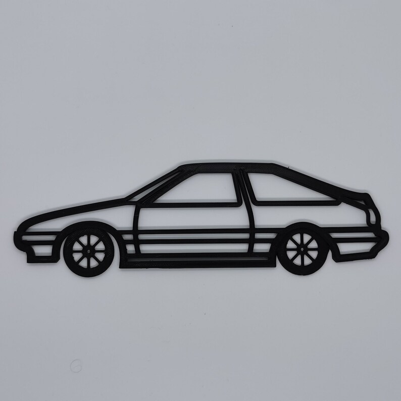 May include: Black outline of a classic sports car, featuring a two-door coupe design. The car has detailed wheels and a sleek profile, set against a plain white background. The design is minimalist and graphic.