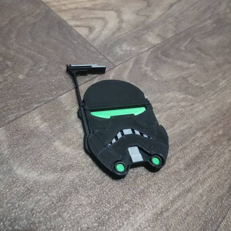 May include: A black and green 3D printed Star Wars Stormtrooper helmet with a raised design. The helmet has a green visor and green eyes.