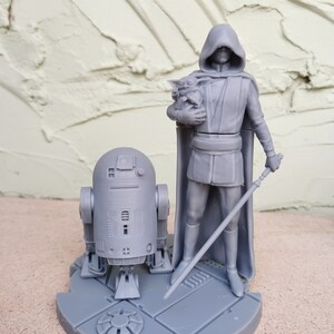 May include: A gray 3D printed figurine set featuring a hooded figure holding a small creature, a droid, and a lightsaber. The figures are standing on a circular base.
