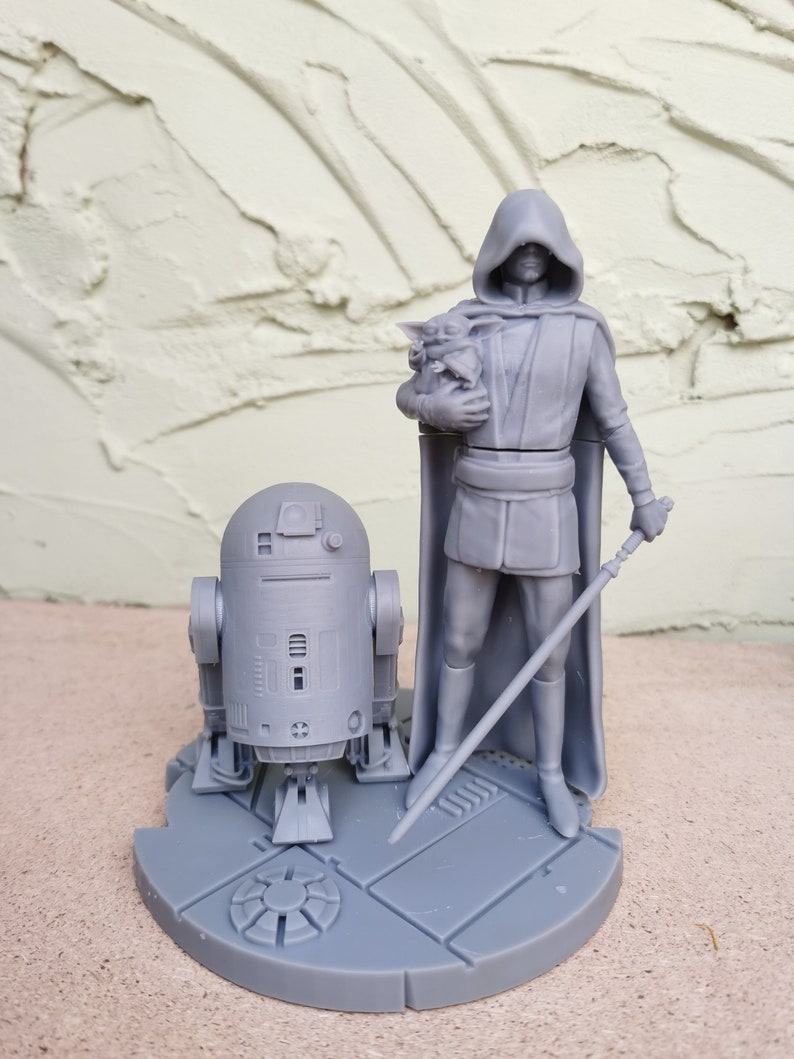 May include: A gray 3D printed figurine set featuring a hooded figure holding a small creature, a droid, and a lightsaber. The figures are standing on a circular base.