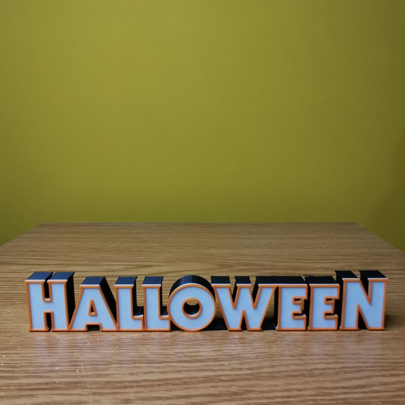 K&ouml;nnte beinhalten: Ein schwarz-wei&szlig;es Halloween-Schild mit orangefarbener Beschriftung. Das Schild zeigt den Text "HALLOWEEN" und steht auf einer Holzoberfl&auml;che.