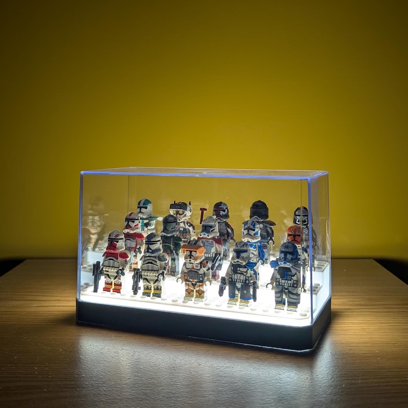 May include: A clear plastic display case with a white LED light base, showcasing a collection of 16 Star Wars clone trooper minifigures. The figures are arranged in a row, with some facing forward and others facing sideways. The figures are all different colors and have various weapons and accessories.