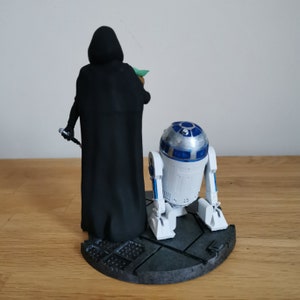 May include: A black-cloaked figure with a lightsaber stands next to a white and blue R2-D2 robot on a gray and black base. The figure is holding a small green figure in its right hand.
