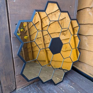 May include: A black framed mirror with a honeycomb design. The mirror is made up of many small hexagonal mirrors that are arranged in a circular pattern. The mirror is reflecting a wooden wall.