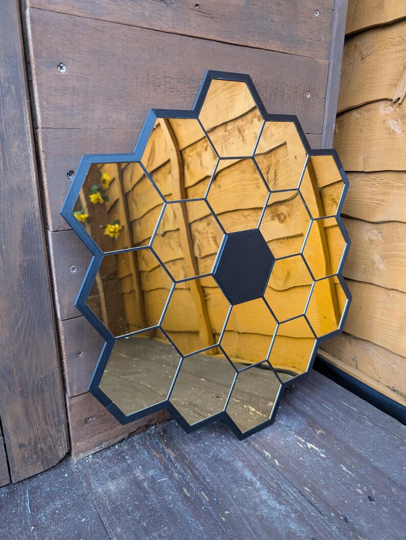 May include: A black framed mirror with a honeycomb design. The mirror is made up of many small hexagonal mirrors that are arranged in a circular pattern. The mirror is reflecting a wooden wall.