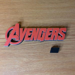 May include: Red and black Avengers logo with a small black rectangular piece. The logo is a stylized wordmark with a red "A" and the word "Avengers" in red with a black outline. The items are on a wooden surface.