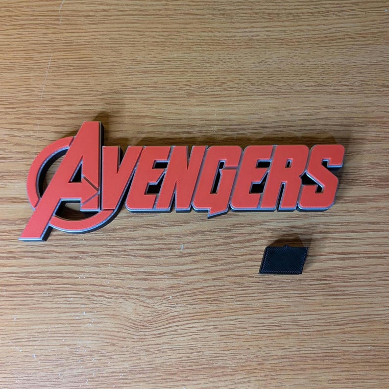 May include: Red and black Avengers logo with a small black rectangular piece. The logo is a stylized wordmark with a red "A" and the word "Avengers" in red with a black outline. The items are on a wooden surface.